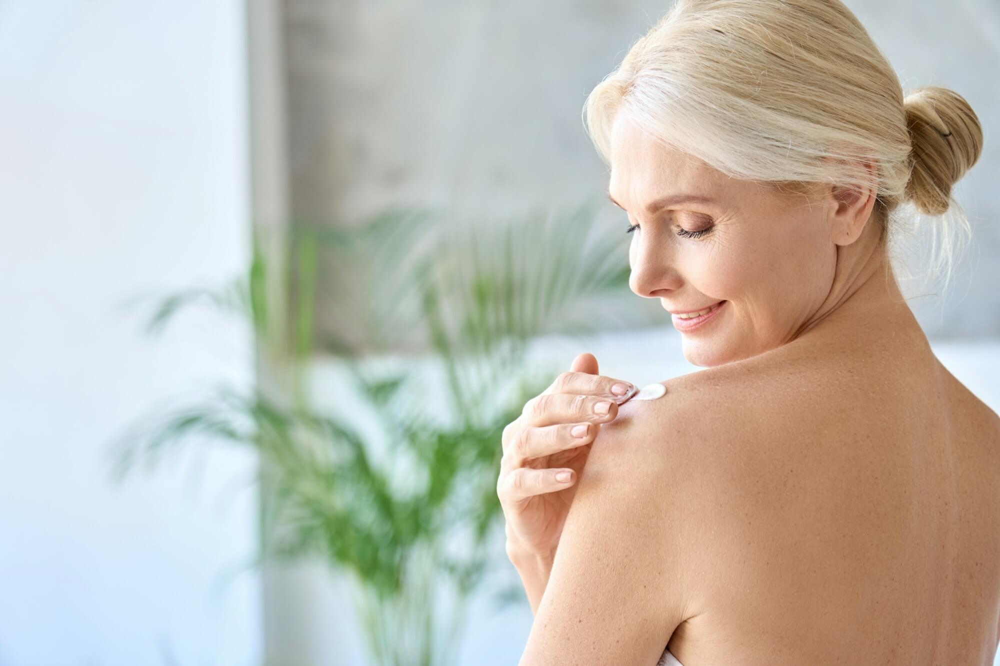 Back view of mid age woman applying body cream. Anti age skincare concept.